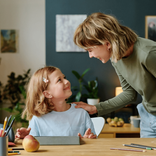 Woman bending over a young girl at a classroom-style table, smiling as they work on school projects with pencils and books nearby.