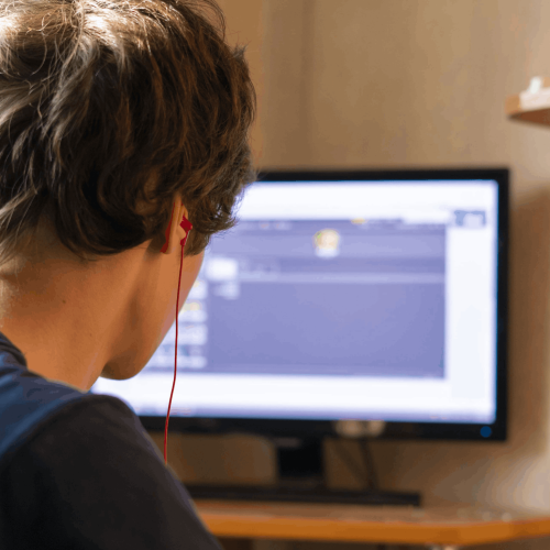 Back view of a person with short brown hair wearing red earbuds, sitting at a desk and looking at a computer monitor in a home/office setup.